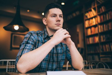 Düşünceli erkek öğrenci şık günlük kıyafetler giyip eğitim sırasında düşünceli bakışlar atıyor. Not defteriyle masada oturup ders çalışmak için zaman harcayan düşünceli beyaz bir öğrenci.