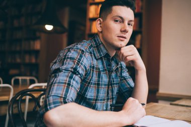 Kareli gömlekli ciddi bir adam kütüphanede masaya oturmuş kameraya bakarken çenesine dokunuyor.