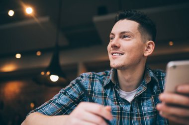 Neşeli siyah saçlı genç adam. Kalem ve akıllı telefonuyla gülümsüyor. Gözlerini kaçırıyor ve lamba ışığında rahat bir kafede oturuyor.