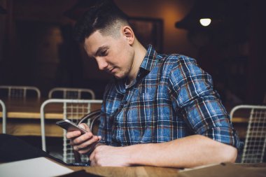 Mavi kareli gömlekli genç adam karanlık odada oturmuş akıllı telefonuyla internette sörf yapıyor.