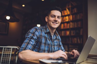 Kütüphanede masa başında oturmuş proje üzerinde çalışırken ve kameraya bakarken rahat gömlekli pozitifsin.