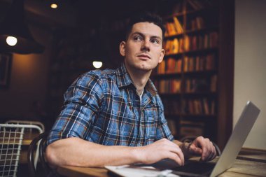 Akıllı günlük gömlekli düşünceli bir erkek öğrenci eğitim için modern dizüstü bilgisayarla masa başında oturuyor ve üniversite kütüphanesinde sınav hazırlıkları sırasında serbest yazar hakkında kafa yoruyor.