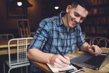 Kafkasyalı mutlu erkek öğrenci e-öğrenim ve araştırma bilgilendirme notlarıyla vakit geçiriyor. Modern dijital netbook ile masaüstünde oturan proje fikirleri üzerinde uzaktan çalışan zeki serbest yazar.