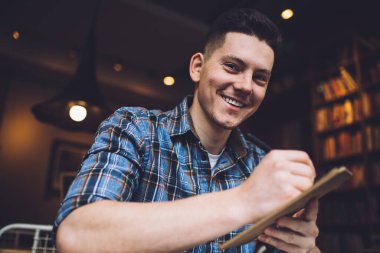 Kütüphanede oturmuş kameraya gülümserken panoya not alan mutlu, genç bir erkek.