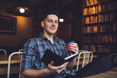 Kamu kütüphanesinde sınav hazırlıkları sırasında, elinde tek kullanımlık yemek kupası olan ve elinde eğitim bilgisini analiz etmek için bilgi içeren bir edebiyat kitabı bulunan erkek öğrencinin portresi.