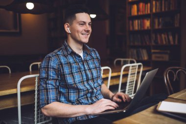 Elektronik kulaklıklı mutlu beyaz adam uzaklardan eğleniyor. Halk kütüphanesinde öğrenim görüyor. Bluetooth kulaklıklı kaygısız erkek öğrenci, dizüstü bilgisayar uygulaması kullanarak müzik dinliyor.