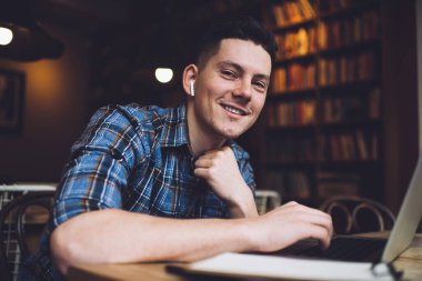 Mavi gömlekli, kablosuz kulaklıklı, dizüstü bilgisayarda sörf yapan kütüphane projesinde çalışan ve kameraya bakan mutlu bir adam.