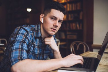 Elektronik kulaklıklı genç hipster adamın portresi, ses kitabı dinlemek için dizüstü bilgisayardaki bluetooth uygulamasına bağlanırken kameraya bakıyor.