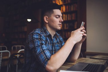 Cep telefonundan mesaj gönderirken kütüphanede elinde netbook ile oturan mavi kareli gömlekli, konsantre genç siyah saçlı adam.