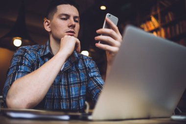 Masada dizüstü bilgisayarla oturup cep telefonuyla internette sörf yaparken çenesine dokunan tişörtlü ciddi bir gencin alçak açısı.