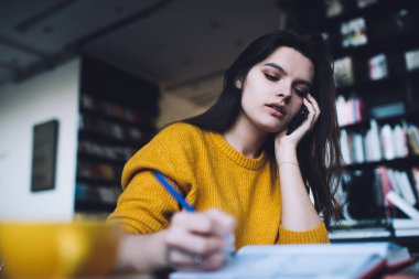 Kafe masasında bir fincan kahveyle otururken cep telefonuyla konuşan ve not defterine yazan gündelik kıyafetlerle ciddi bir genç kadının alçak açısı.