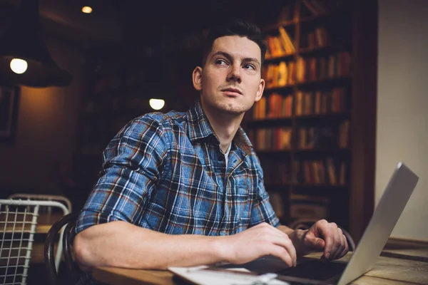 Akıllı günlük gömlekli düşünceli bir erkek öğrenci eğitim için modern dizüstü bilgisayarla masa başında oturuyor ve üniversite kütüphanesinde sınav hazırlıkları sırasında serbest yazar hakkında kafa yoruyor.