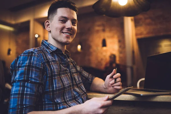 From below side view of positive young guy in plaid shirt using netbook and smartphone with stylus while working remotely sitting in cozy cafe