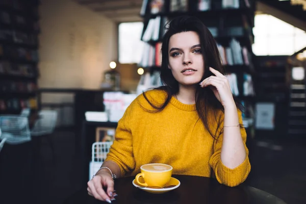 在现代咖啡馆喝咖啡时 身穿黄色毛衣的年轻女士坐在桌旁 抚摸着头发 — 图库照片