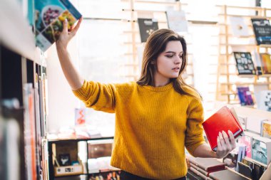 Günümüz güneşli kütüphanesinde boş zaman geçirirken kitaplıkların yanında duran ve kitapları seçen gündelik kıyafet giyen pozitif zeki bayan öğrenci.