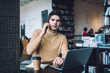 Modern çalışma sahasında internet rehberi ve bir fincan kahveyle uzaktan iş yaparken kameraya bakarak odaklanmış genç bir adam.