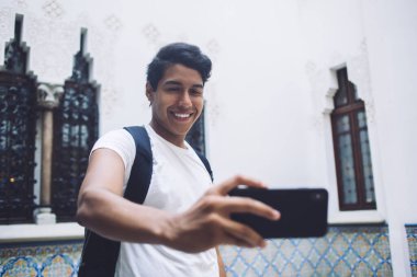 Komik Latin hipster adam selfie görüntülerine tıklıyor. Akıllı telefon kamerasına gülümsüyor. Neşeli erkek oyuncu 20 yaşında.