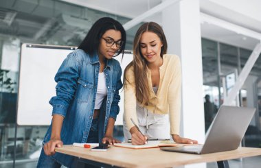 Avrupalı iş kadını siyah iş arkadaşına not defterine bir şey yazıyor. Modern başarılı kadın kavramı. Takım çalışmasının bir fikri. Ofiste dijital cihazları ve işaretleri olan genç kızlar