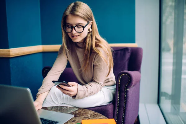 カジュアルなセーターや流行の眼鏡を着た若い女性は 現代の紫色のアームチェアに座って スマートフォンを使用している間 ラップトップ上でインターネットを閲覧 — ストック写真
