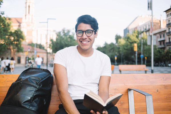 Portrait of cheerful male tourist with backpack holding textbook planner with checklist for visiting, happy Latino student with education notepad or literature best seller in hand smiling at camera