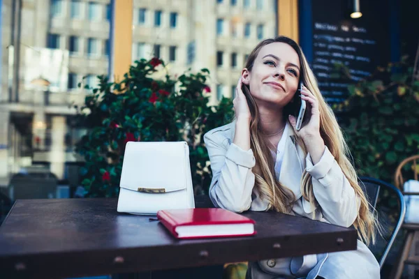 穿着时髦外套的快乐的年轻女生坐在桌旁 咖啡店里拿着白色皮包和笔记本 一边看着一边打电话 — 图库照片