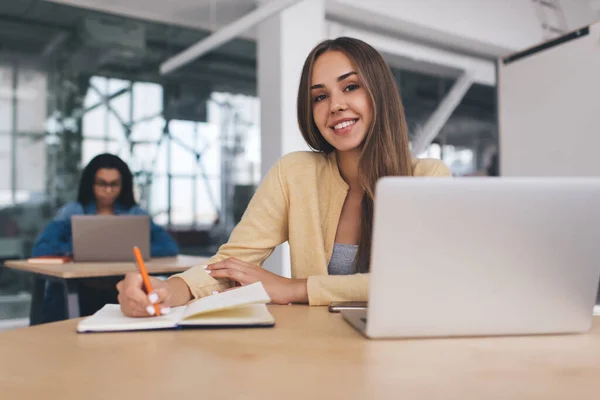 微笑的欧洲裔女商人在笔记本上写东西 而她的欧洲裔女同事在笔记本电脑上做背景工作 现代成功女性的概念 坐在办公室办公桌前的女孩 — 图库照片
