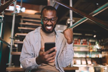 Koyu tenli erkek blogcu ağ hedefini web popülerliğiyle kutlarken cep telefonunda 4G kablosuz ağ kullanıyor. Akıllı telefon aygıtındaki e-posta mesajından memnun olan neşeli adam.