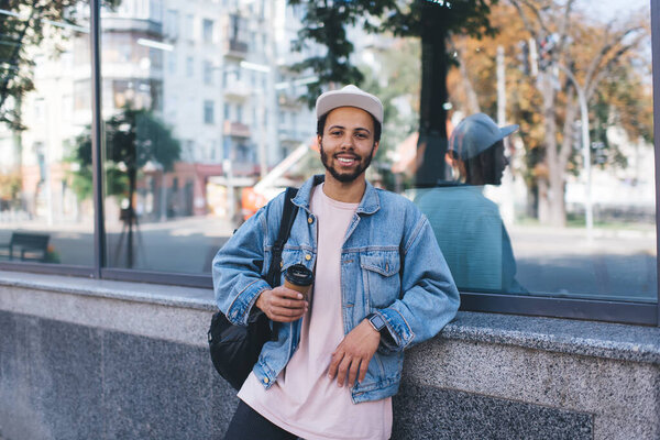 Half length portrait of carefree hipster guy in trendy denim clothes smiling at camera during coffee break in touristic city, happy traveller in cap holding takeaway caffeine beverage and posing