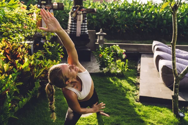 在度假胜地的绿地草坪上练习瑜伽时 穿着白色上衣站在伸展的侧角姿势的快乐女性 — 图库照片