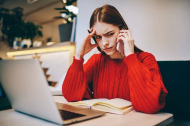 Kırmızı kazaklı, ciddi bir kadın akıllı telefonla konuşurken dizüstü bilgisayar ve modern kafede notlar alıyor.