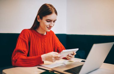 Uzun saçlı, gülümseyen, kırmızı örgü kazaklı serbest çalışan, tablet kullanan ve modern çalışma alanındaki projeye uzaktan bakarken ekrana bakan bir kadın.