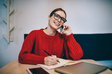 Kırmızı örgü kazak ve gözlüklü serbest çalışan mutlu bir kadın. Not defterine not alıyor, akıllı telefonu sohbet için kullanıyor ve yukarı bakıyor.