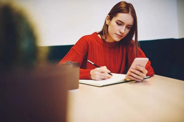 カフェでスマホを使いながら テーブルに座ってノートを書きながら明るい赤いセーターを着た集中学生 — ストック写真