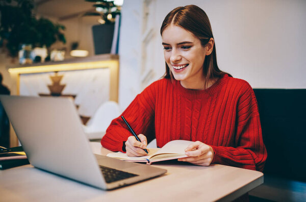 Happy youthful brunette wearing warm sweater looking at laptop screen and writing thoughts in notebook while sitting at wooden table in light cafeteria