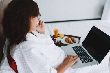 Kuala Lumpur 'da otel odasında dizüstü bilgisayarla yatarken cep telefonuyla konuşan, beyaz bornozlu kimliği belirsiz bir kadın.