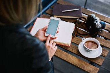 Kafeteryadaki masasında oturan yüzü olmayan kadının yüksek açılı görüntüsü. Elinde çay, elinde kalem, fotoğraf makinesi ve kitapla telefonu kontrol ederken.