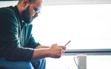 Dikkate değer sakallı, günlük kıyafetler giymiş, gözlüklü, dinlenirken internette sörf yapan ve kahve molası veren erkeklerin yan görüntüsü.