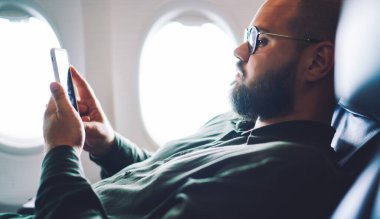 Pencere kenarındaki rahat koltukta oturan ve gündüz gözlüklü akıllı telefon kullanan yetişkin bir erkek.