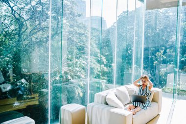 Heyecanlı, gözlüklü, internette gezinen, el ele tutuşan ve iş yerinde gün ışığında camdan duvarları olan ekrana bakan uzaktan kumandalı kadın.