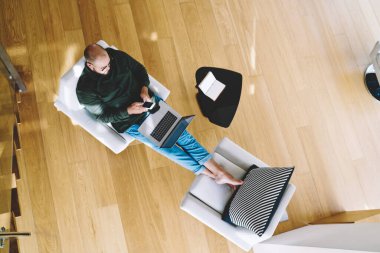 Yukarıdan, gözlüklü, cep telefonlu, dizleri dizlerinde dizleriyle koltukta oturan uzaktan kumandalı bir erkek.