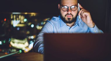 Ekin yoğunlaştırılmış gözlüklü erkek serbest çalışan masada oturmuş bilgisayarını kullanarak geceleri cep telefonuyla konuşuyor.