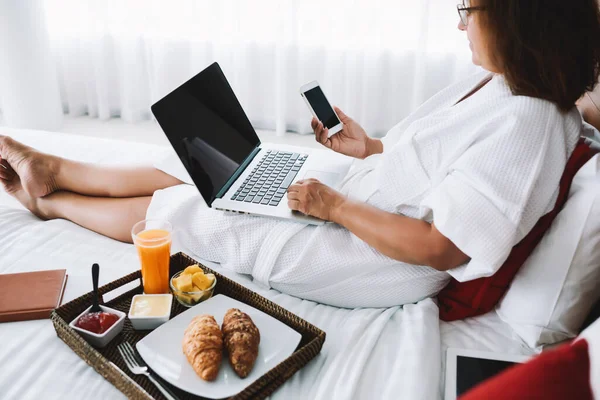 Beyaz sabahlık giymiş, beyaz elbiseli, akıllı telefonuyla mesajlaşan ve Kuala Lumpur 'da dizüstü bilgisayarla yatakta dinlenen bir kadının yan görüntüsü.