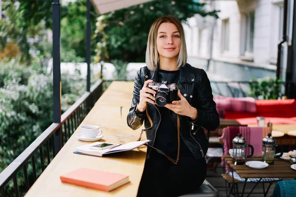 カメラを手にした現代的な通りの食堂に立つ若い女性とテーブルでドリンクやノートパソコンを持ち カメラを見て — ストック写真