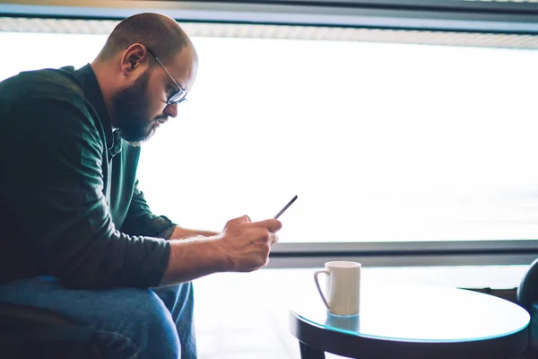 聚精会神 蓄着胡子的男性坐在圆桌边喝咖啡 同时利用智能手机享受闲暇的侧视图 — 图库照片