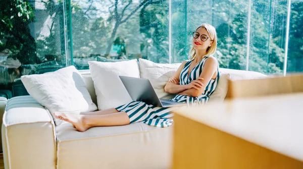 阳光灿烂的现代玻璃阳台上 年轻快乐的女士躺在舒适的沙发上 抱着垫子 在笔记本电脑上观看视频的侧视图 — 图库照片