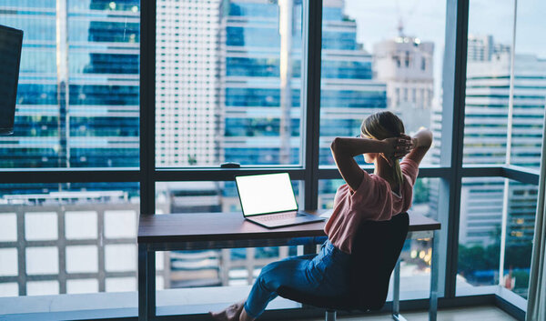 Вид сзади женщины в повседневной одежде, сидящей за столом с ноутбуком и руками, сжатыми за головой во время перерыва на работе