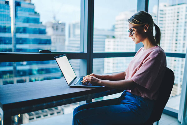 Боковой вид довольной женщины-предпринимателя в повседневной одежде и очках, внимательно смотрящей на экран ноутбука, работая в современном офисе с окном во всю стену