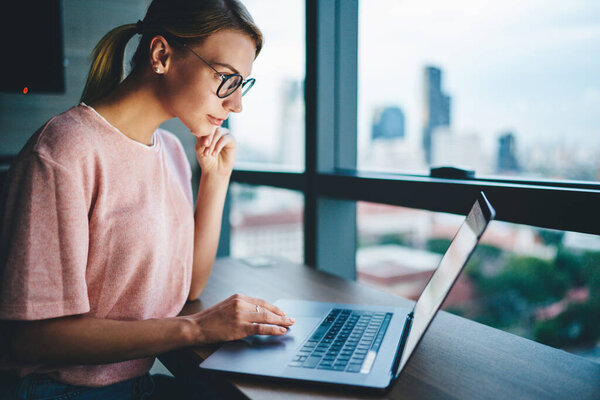 Боковой вид очаровательной женщины в повседневной одежде и очках, наклоняющихся над ноутбуком и смотрящих на экран во время работы над проектом в современном рабочем пространстве вечером