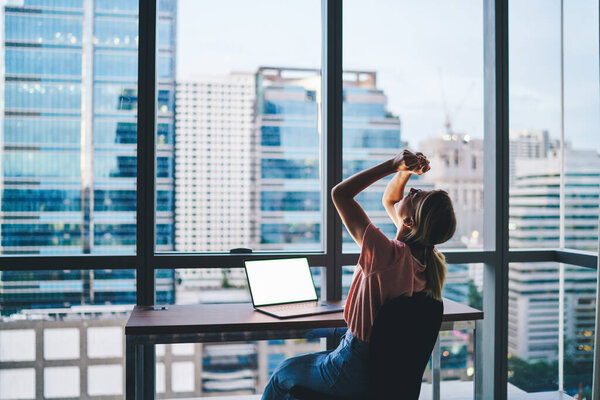 Вид сбоку на молодую работницу в повседневной одежде, сидящую за столом с ноутбуком, смотрящую вверх и сжимающую руки в кулаки во время празднования успеха