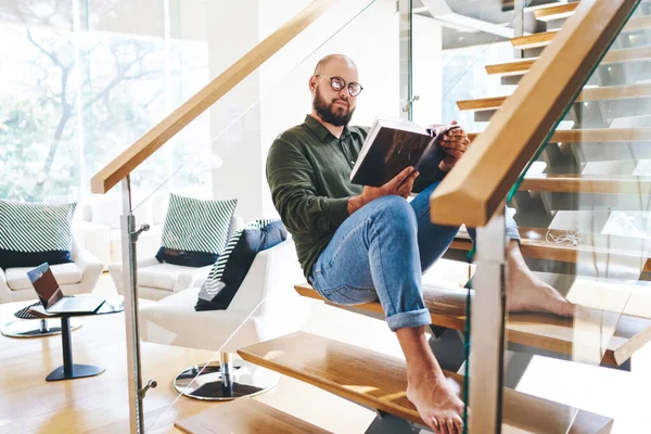 穿着休闲装和眼镜 满头大胡子的男人坐在现代化公寓的楼梯上看书 — 图库照片